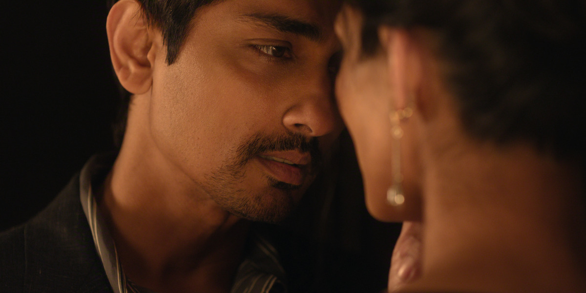 A man and woman in an intimate moment, facing each other closely in a dimly lit setting; the man looks at the woman tenderly while she wears earrings and soft lighting creates a romantic atmosphere.