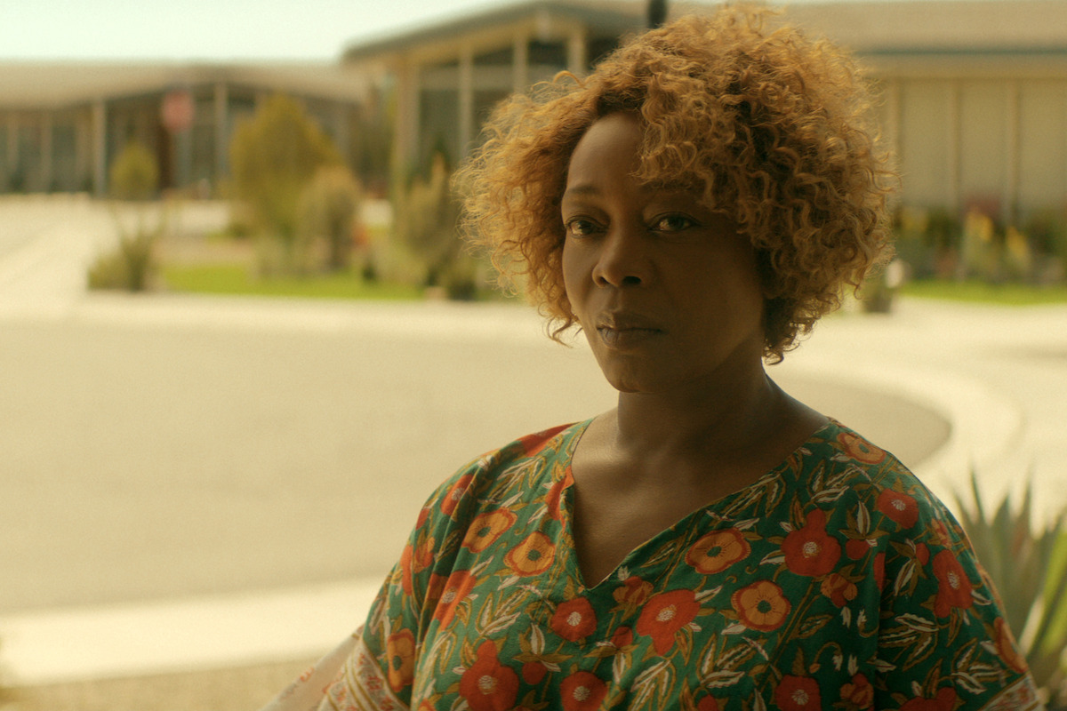 Alfre Woodard as Judy, with curly hair and floral shirt standing outdoors in a sunny courtyard, with modern buildings and greenery in the background.