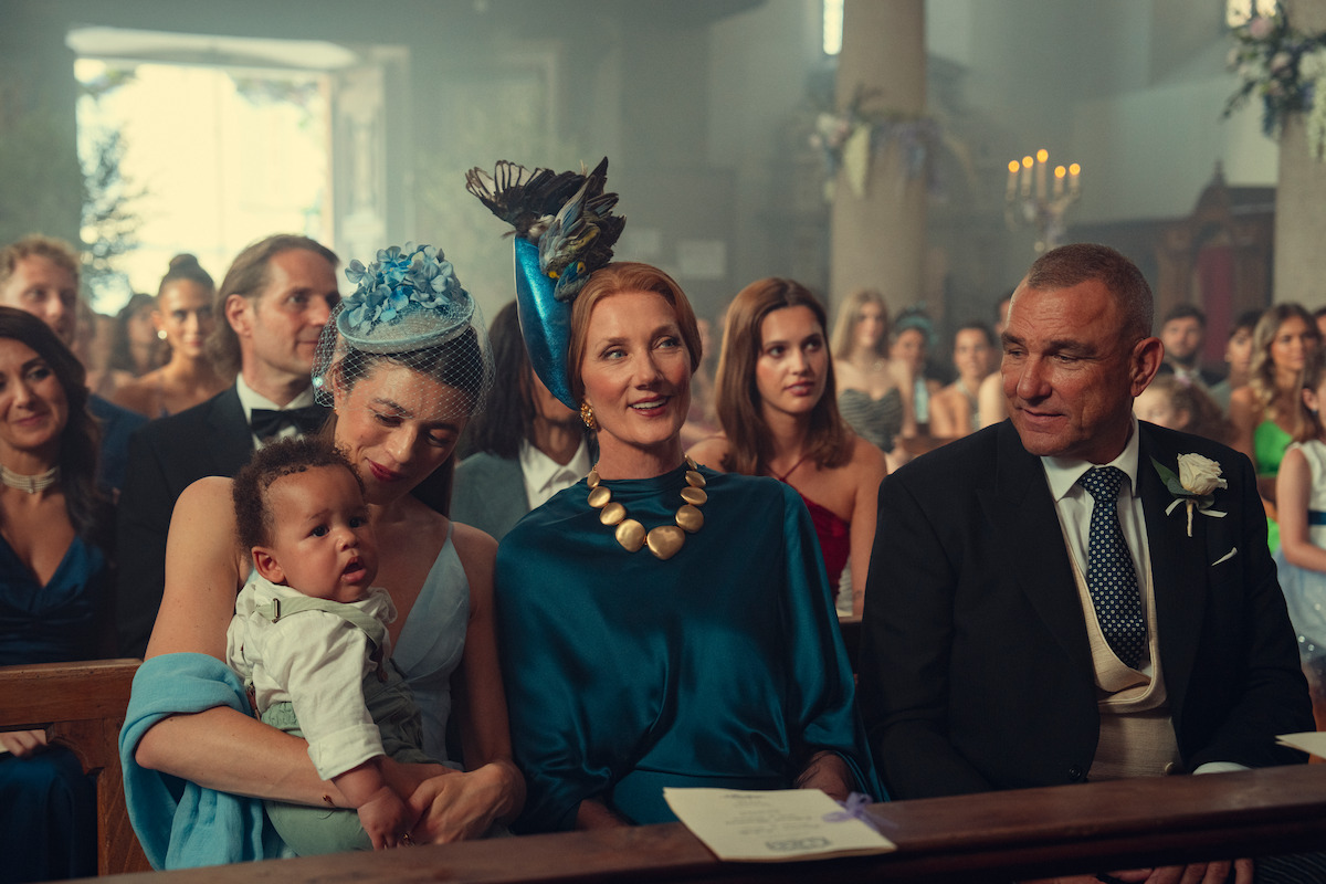 People dressed formally sitting in pews at a wedding ceremony inside a church, with women wearing hats and a woman holding a baby; candles and floral decorations are visible in the background.