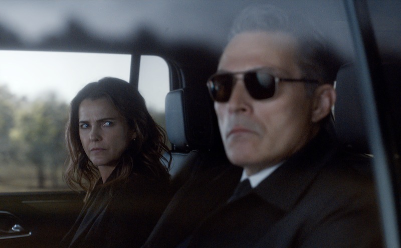 A woman with a serious expression sits in the back seat of a car next to a man wearing dark sunglasses and a suit, both looking out the window.