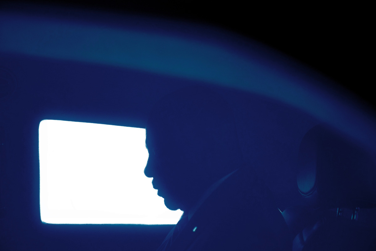 Silhouette of a person sitting inside a dimly lit car, illuminated by bright light from the window. Overall mood is mysterious and contemplative, with deep blue tones adding to the atmospheric setting.