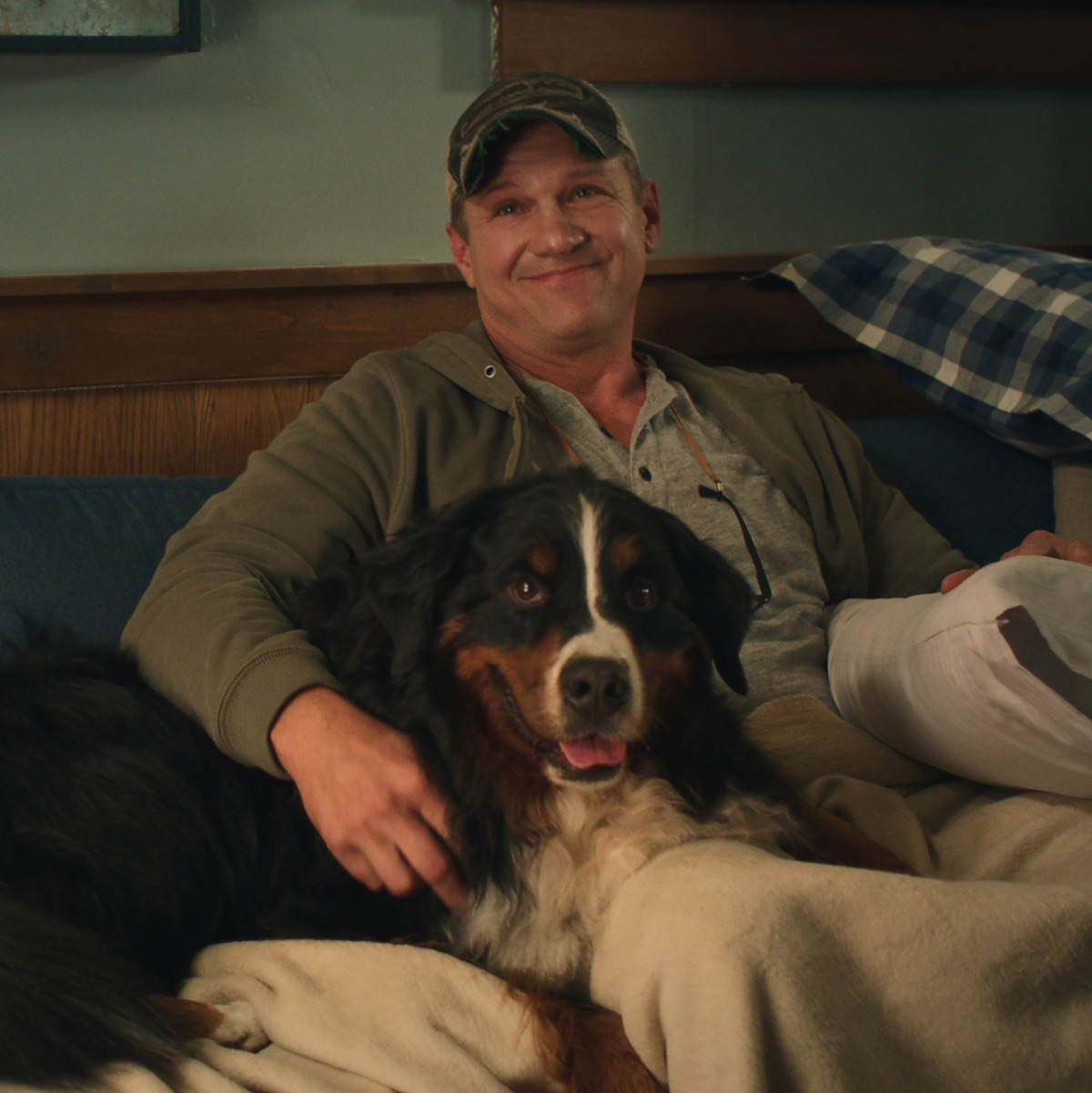 Man sitting on a couch in a cozy room, smiling while relaxing under a blanket with a large Bernese Mountain Dog, warm lighting and plaid pillows in the background.