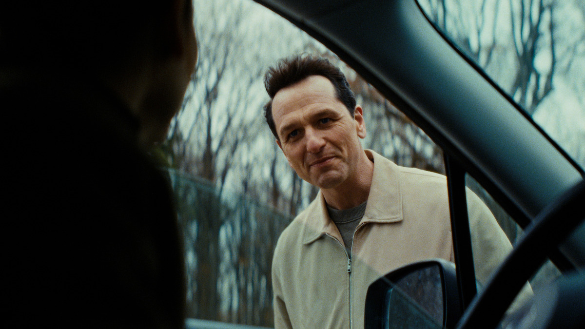 A man in a beige jacket stands outside a car, looking in through the open door. The environment is outdoors with bare trees, suggesting late autumn or winter. The mood appears tense or serious.
