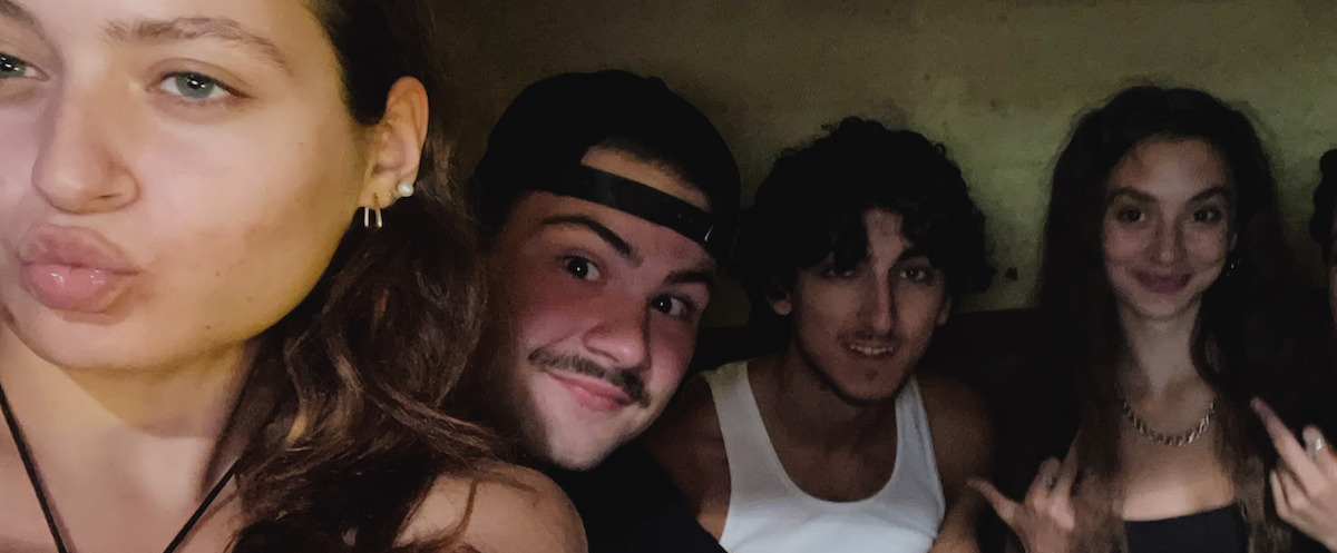 Four young adults pose closely together in a dimly lit indoor setting; one person in the foreground makes a duck face, while the others smile and gesture casually towards the camera, creating a lively and relaxed atmosphere.