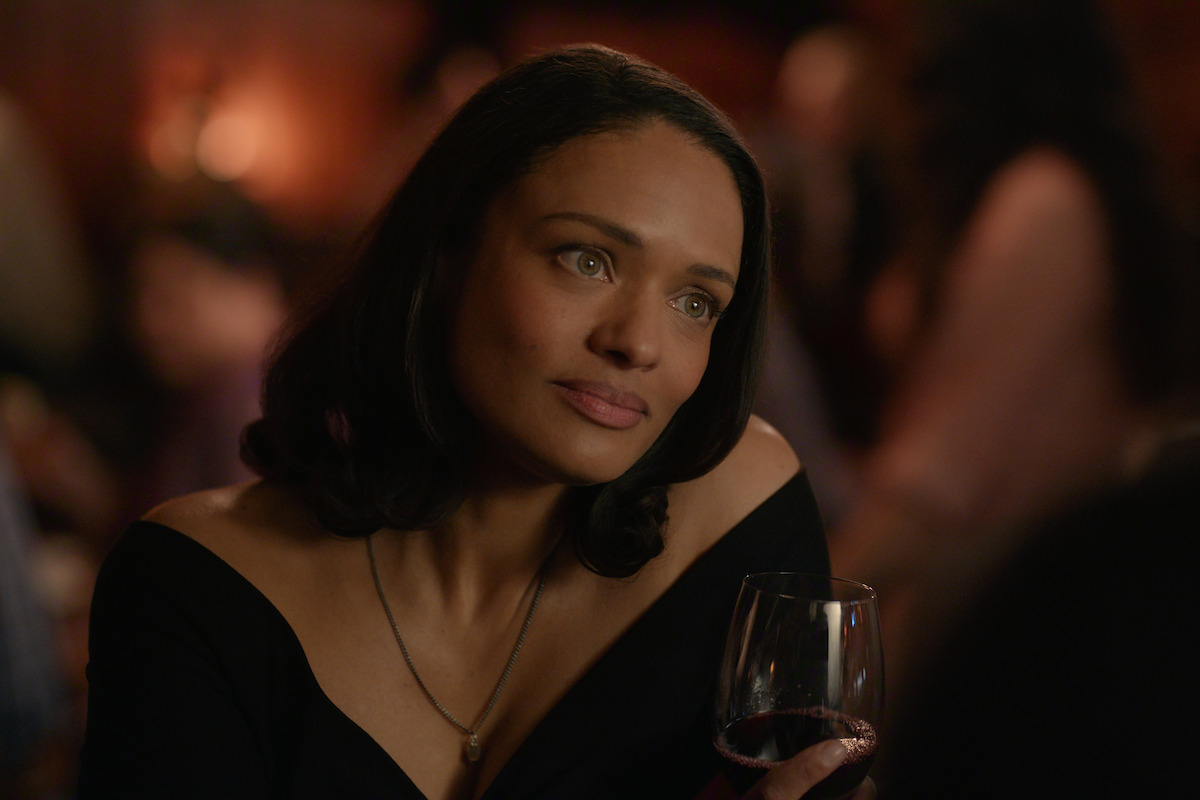 Woman in a black off-shoulder dress holding a glass of red wine, sitting in a dimly lit bar or restaurant, with a thoughtful expression and blurred people in the background.