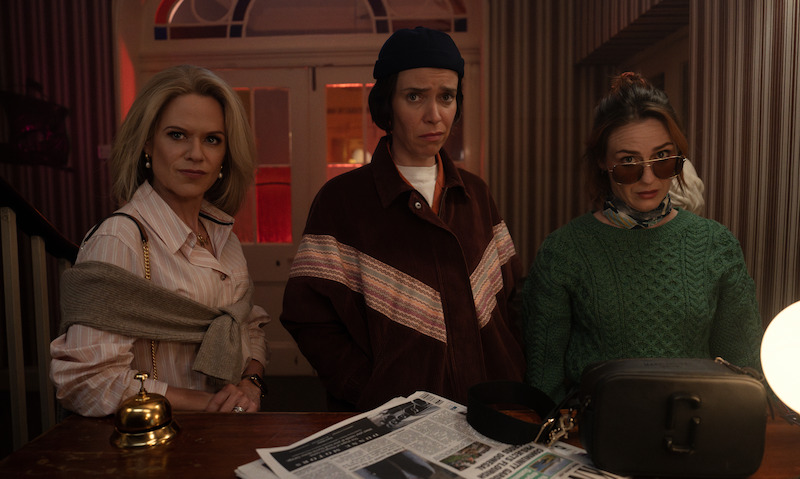 Three women stand at a dimly lit reception desk with a bell, newspaper, and handbags, looking serious and inquisitive in a vintage-styled interior, creating a tense, mysterious mood.