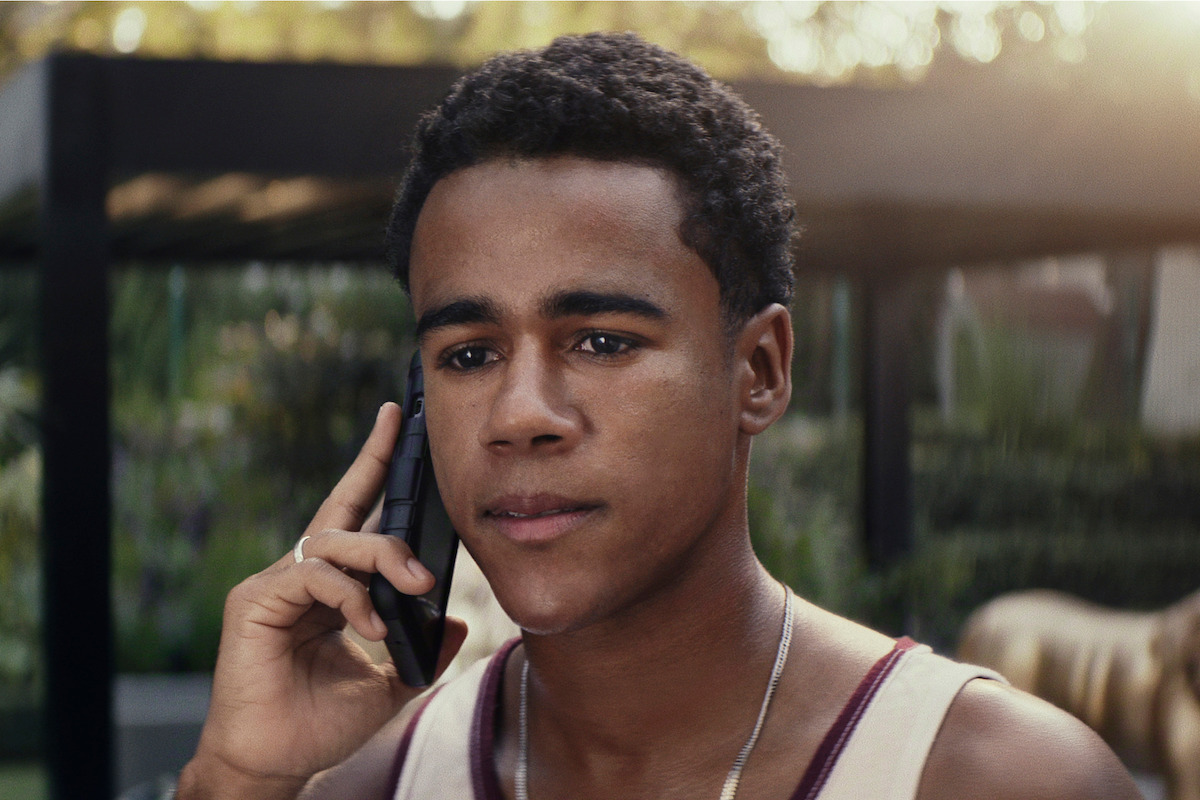 Teenage boy wearing a tank top talks on a smartphone outdoors, with a blurred garden or park background and soft sunlight.