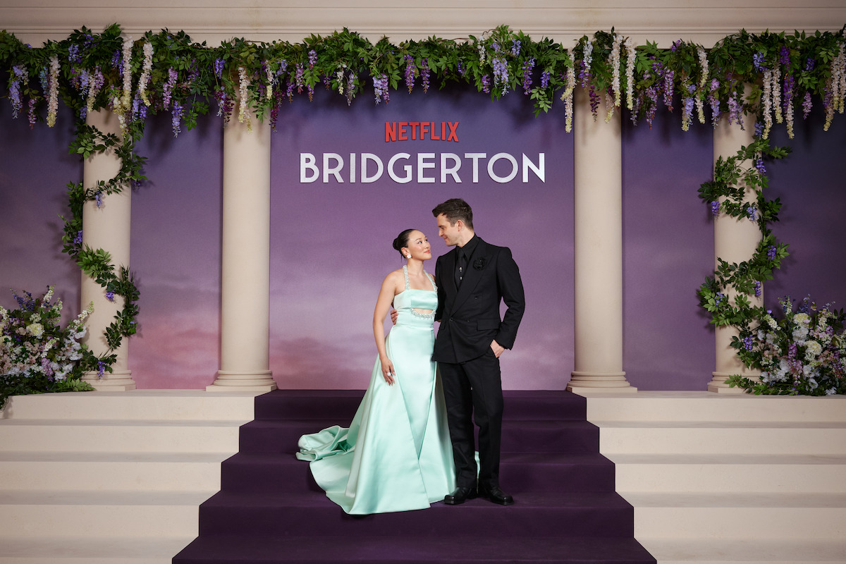 Two people dressed formally stand on purple steps decorated with flowers, posing in front of a Netflix Bridgerton backdrop designed like a grand entrance with columns and floral garlands, suggesting a themed event or premiere.