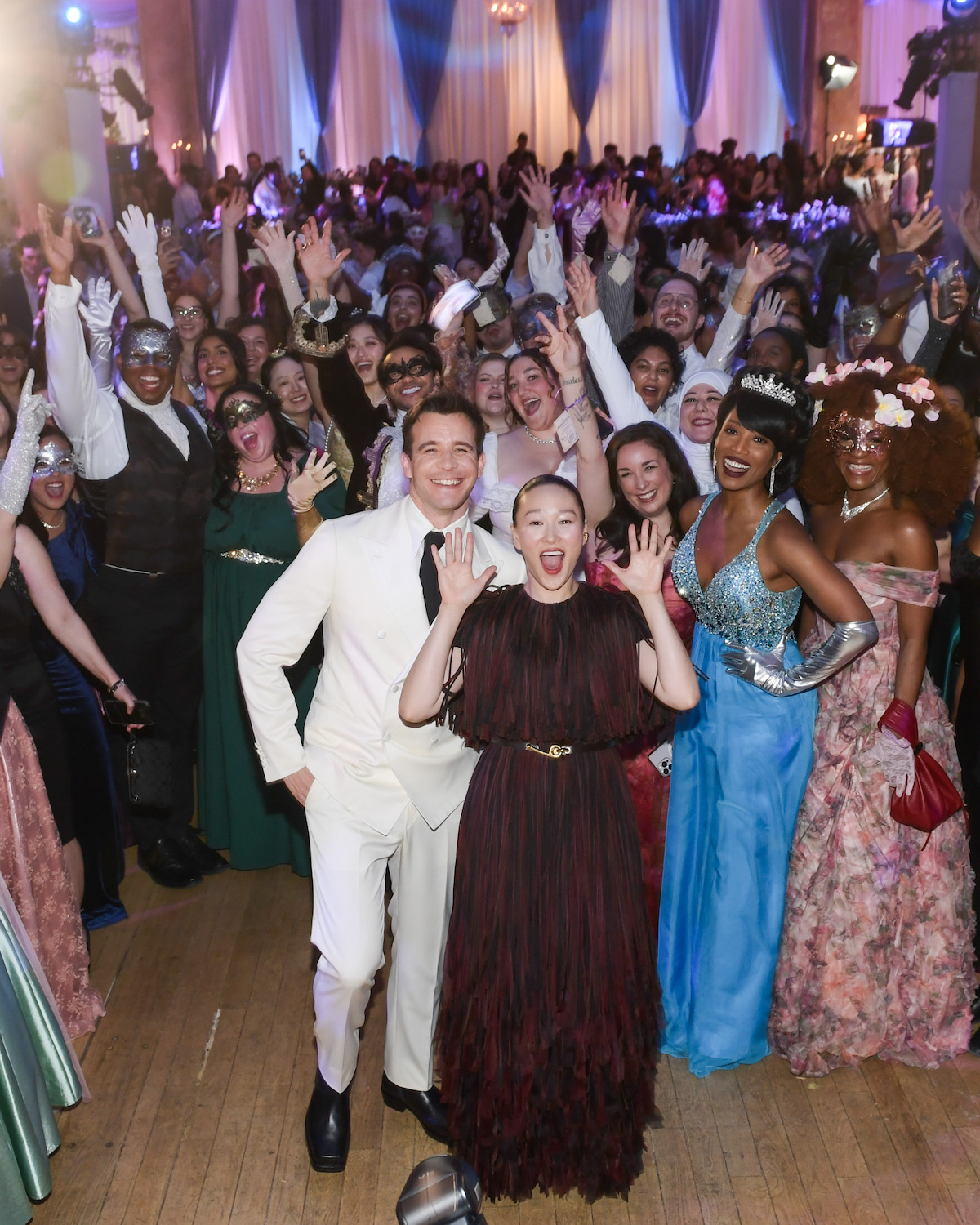 A large group of people in formal attire, many wearing masks, pose and smile excitedly at a festive indoor event with draped curtains and chandeliers, suggesting a celebratory masquerade ball or themed party atmosphere.