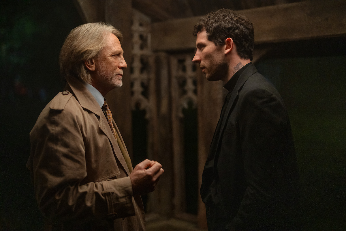 Two men stand face to face in a dimly lit, rustic wooden structure at night, engaged in a serious conversation; one in a trench coat and tie, the other in dark clothes, creating a tense, dramatic atmosphere.