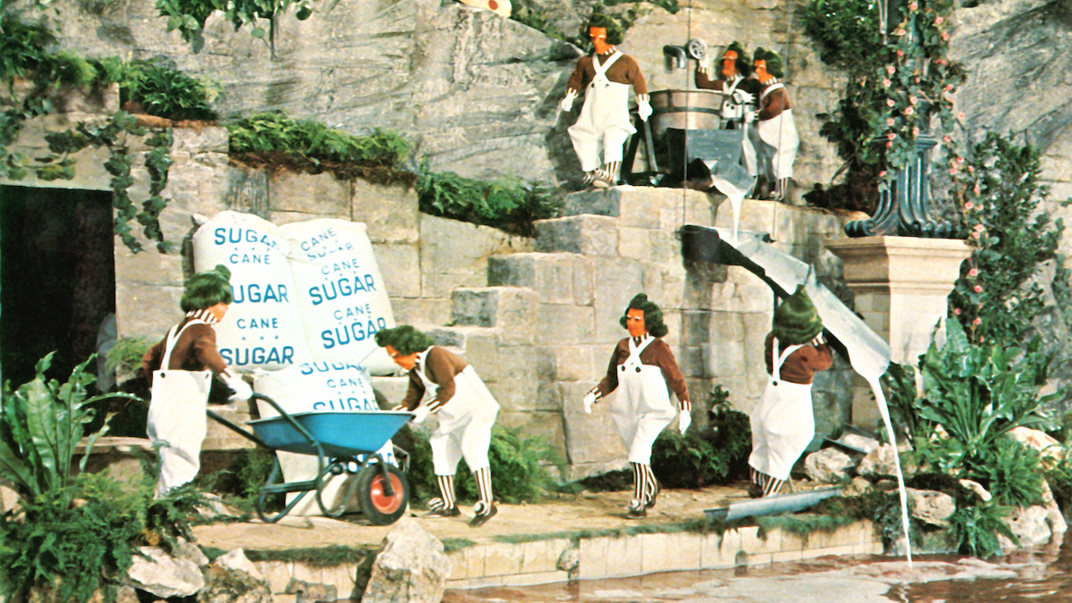 People dressed in costumes work in a whimsical, lush environment carrying large sugar bags and using carts and chutes near stone steps and flowing water, surrounded by greenery and a fantastical set design.