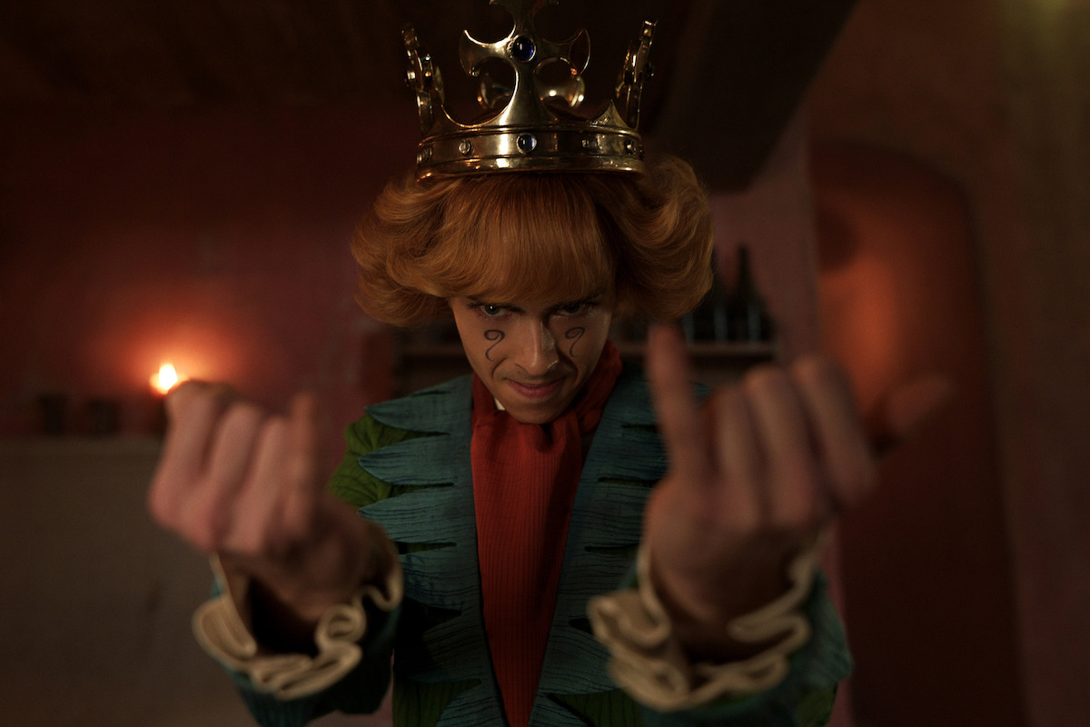 A man with face paint and a gold crown in colorful, ornate clothing gestures dramatically with both hands indoors, in a dimly lit, reddish room with candles and shelves in the background.