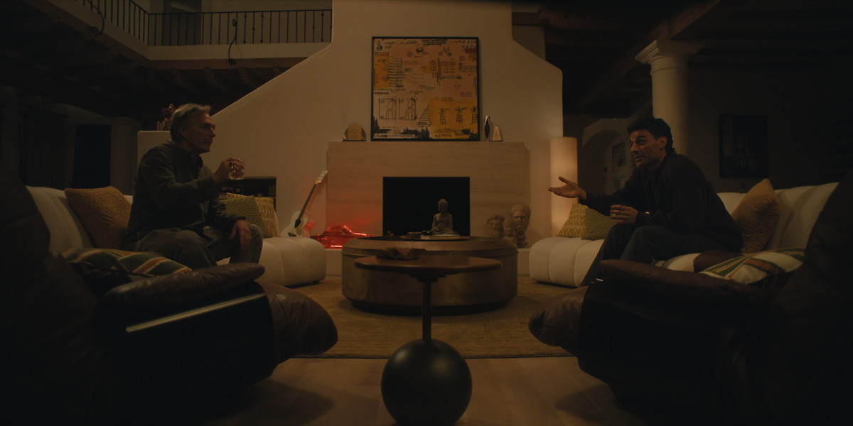 Two men sit facing each other in a dimly lit, cozy living room with modern decor, plush sofas, a round coffee table, fireplace, abstract art, lamps, and various decorative items, engaged in conversation.