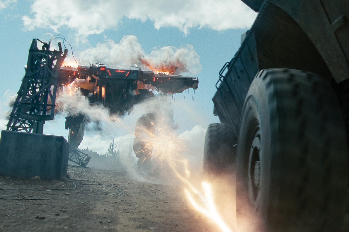 A large, fiery robot shoots lasers at a moving truck on a dusty battlefield under a cloudy sky, with smoke, debris, and intense action in an outdoor, post-apocalyptic environment.