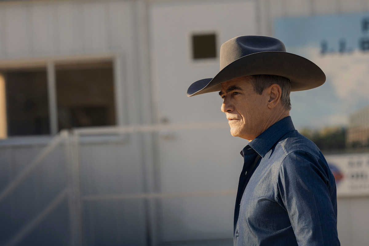 Man wearing cowboy hat and blue shirt standing outdoors in front of a light-colored building with a door and window, appearing to be in a rural or construction site setting during daylight.