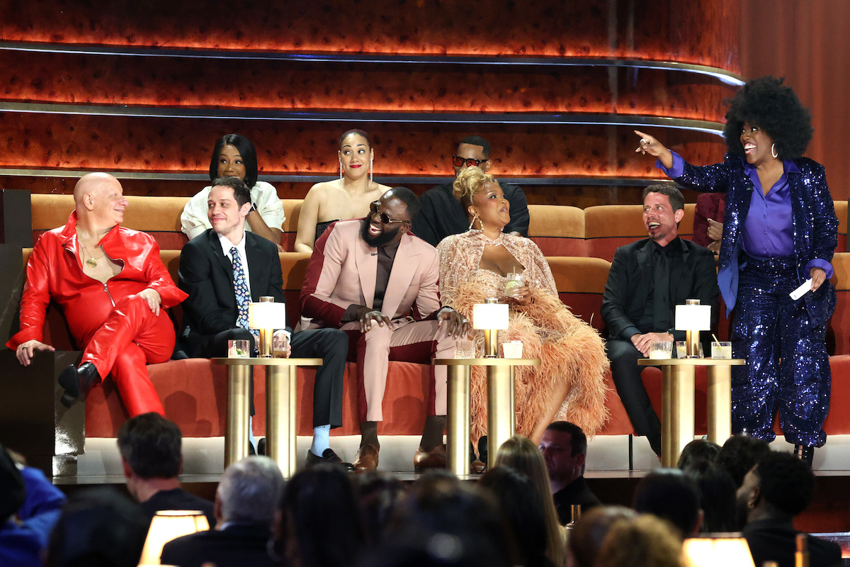 A group of people onstage in a theater setting, dressed in formal and colorful outfits, sitting on plush seats and laughing as a woman in a sparkling suit stands and gestures, with an audience watching in front.