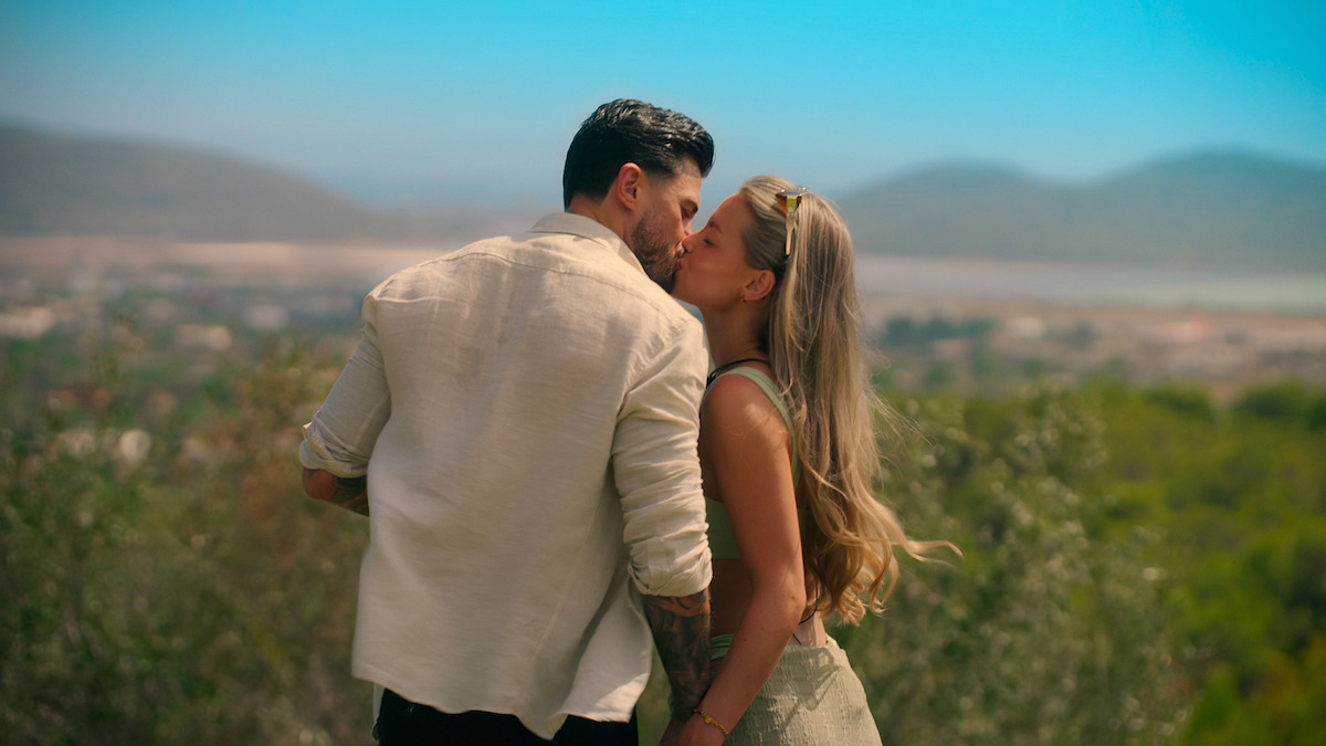 Couple kissing outdoors with scenic mountains and blue sky in the background, surrounded by nature and greenery on a sunny day. Romantic moment in a peaceful natural setting.