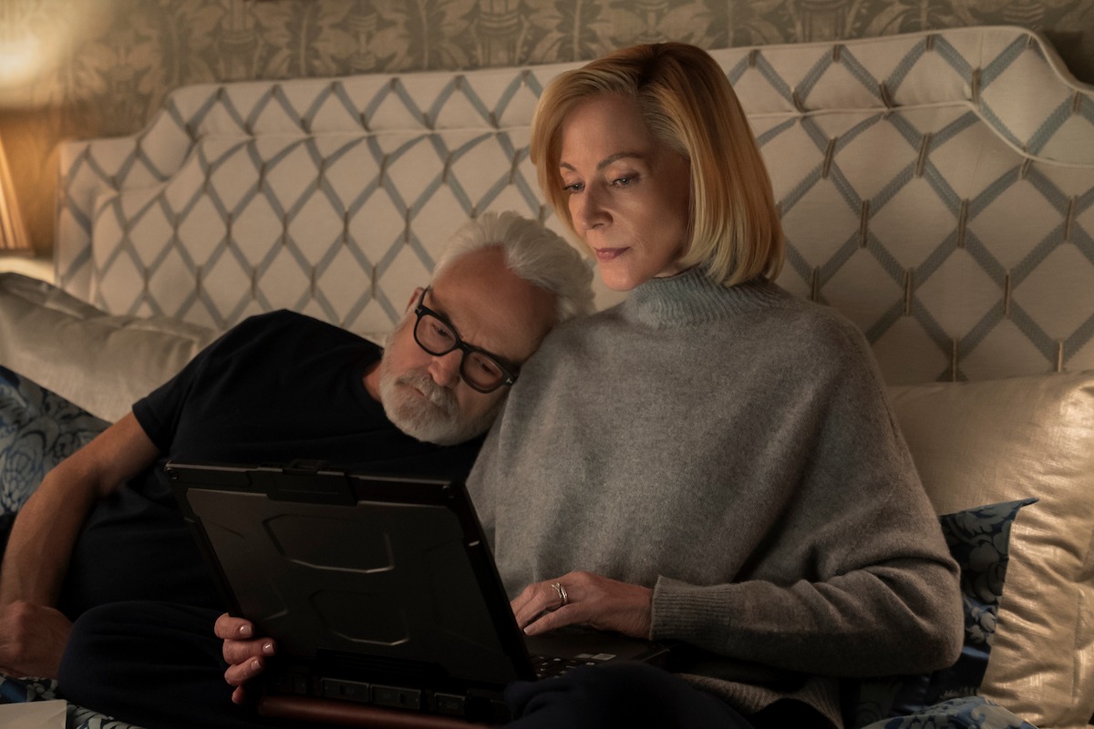 Older couple relaxing in bed together, man resting his head on woman's shoulder while she uses a tablet, cozy bedroom setting with patterned headboard and soft lighting.