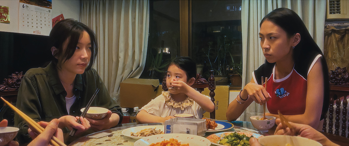Three people, including a young girl, sit around a dining table with food, sharing a meal in a cozy, warmly lit home. The mood is tense or serious, with subdued expressions and focused body language.