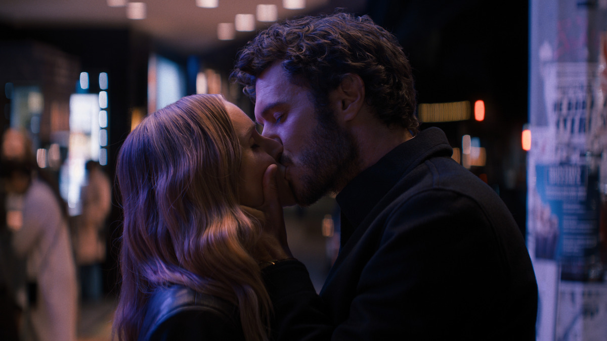 A man and woman share a romantic kiss at night on a city street, surrounded by blurred lights and posters on a wall, creating a moody and intimate urban atmosphere.