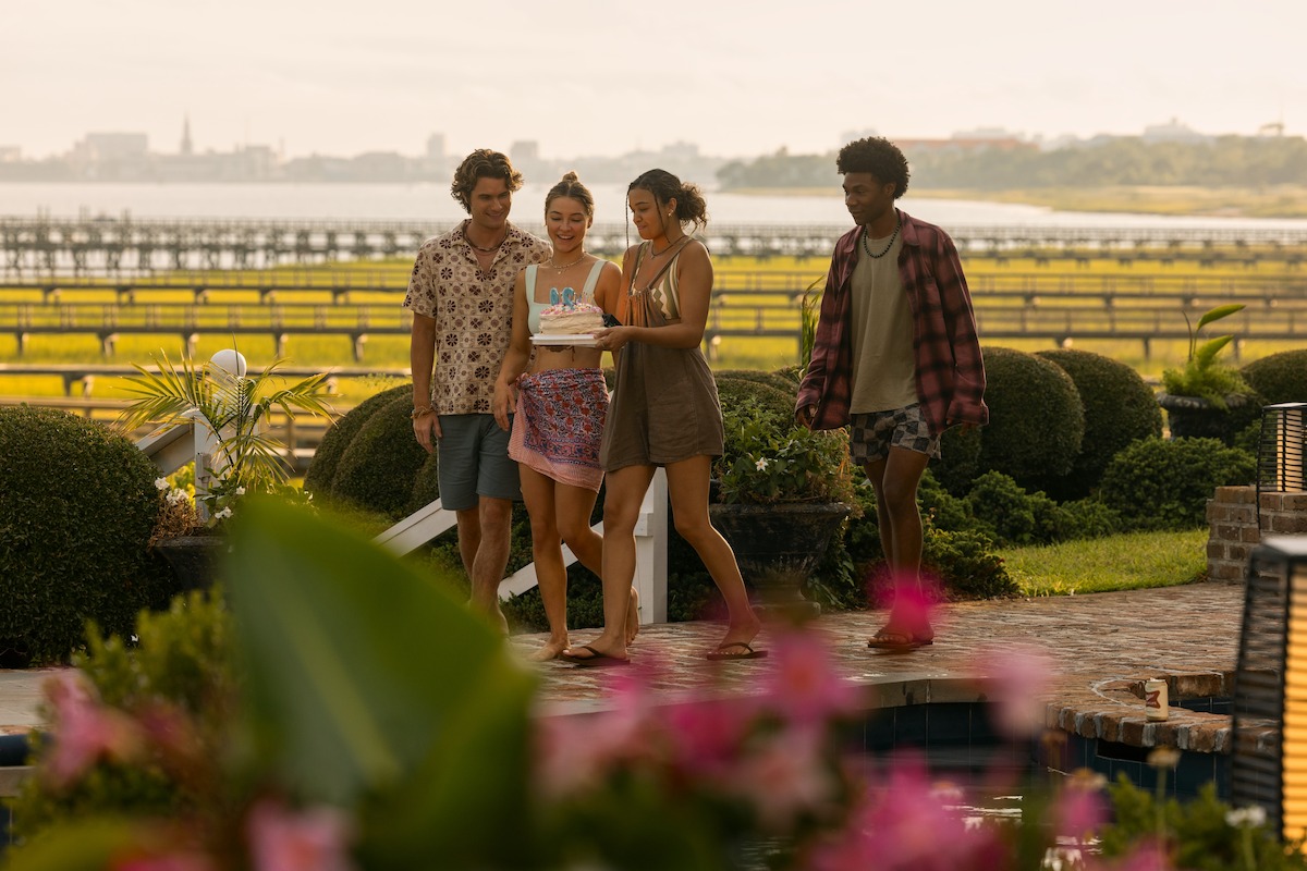 Quatro jovens adultos caminham juntos aproveitando um dia tranquilo de sol, ao lado de uma piscina em um jardim exuberante com arbustos, flores e vista para a água e para a paisagem urbana ao longe.