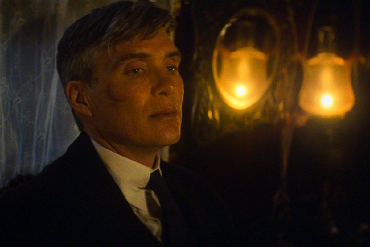 Man in a dark suit, white shirt, tie, and flat cap stands outdoors, facing forward in a moody, dimly lit environment, conveying a serious, intense expression.