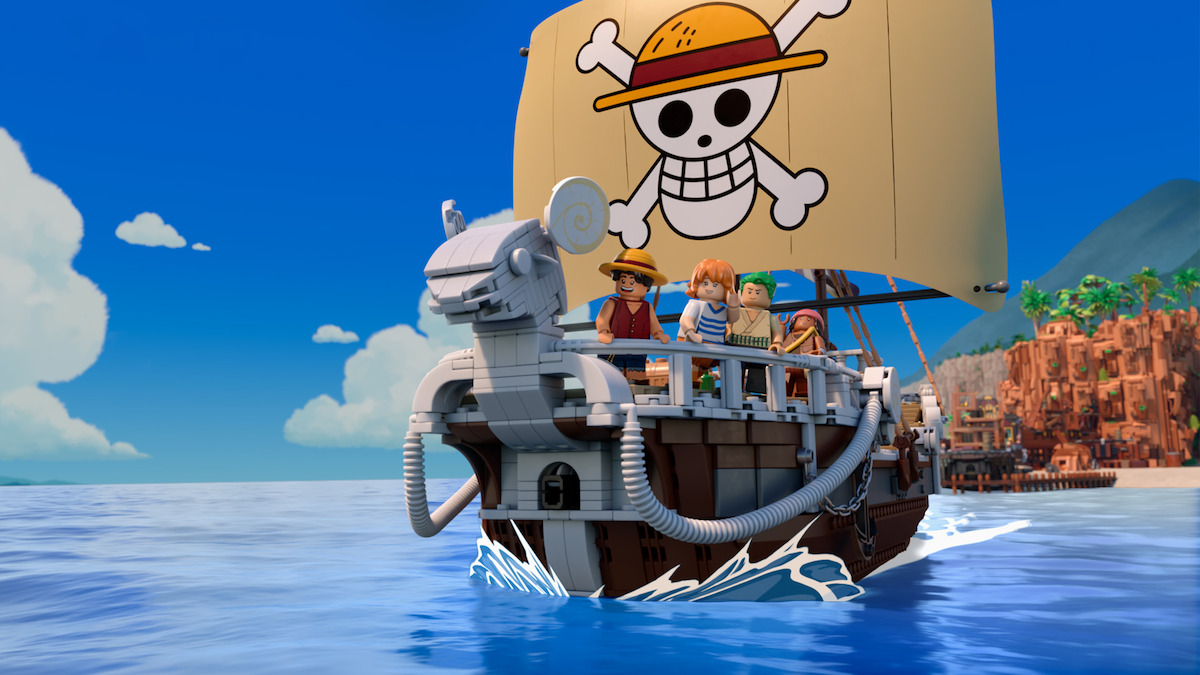 LEGO pirate ship with four minifigures sailing on blue water, featuring a sail with a skull and crossbones wearing a straw hat, under a clear blue sky with a coastline and mountains visible in the background.