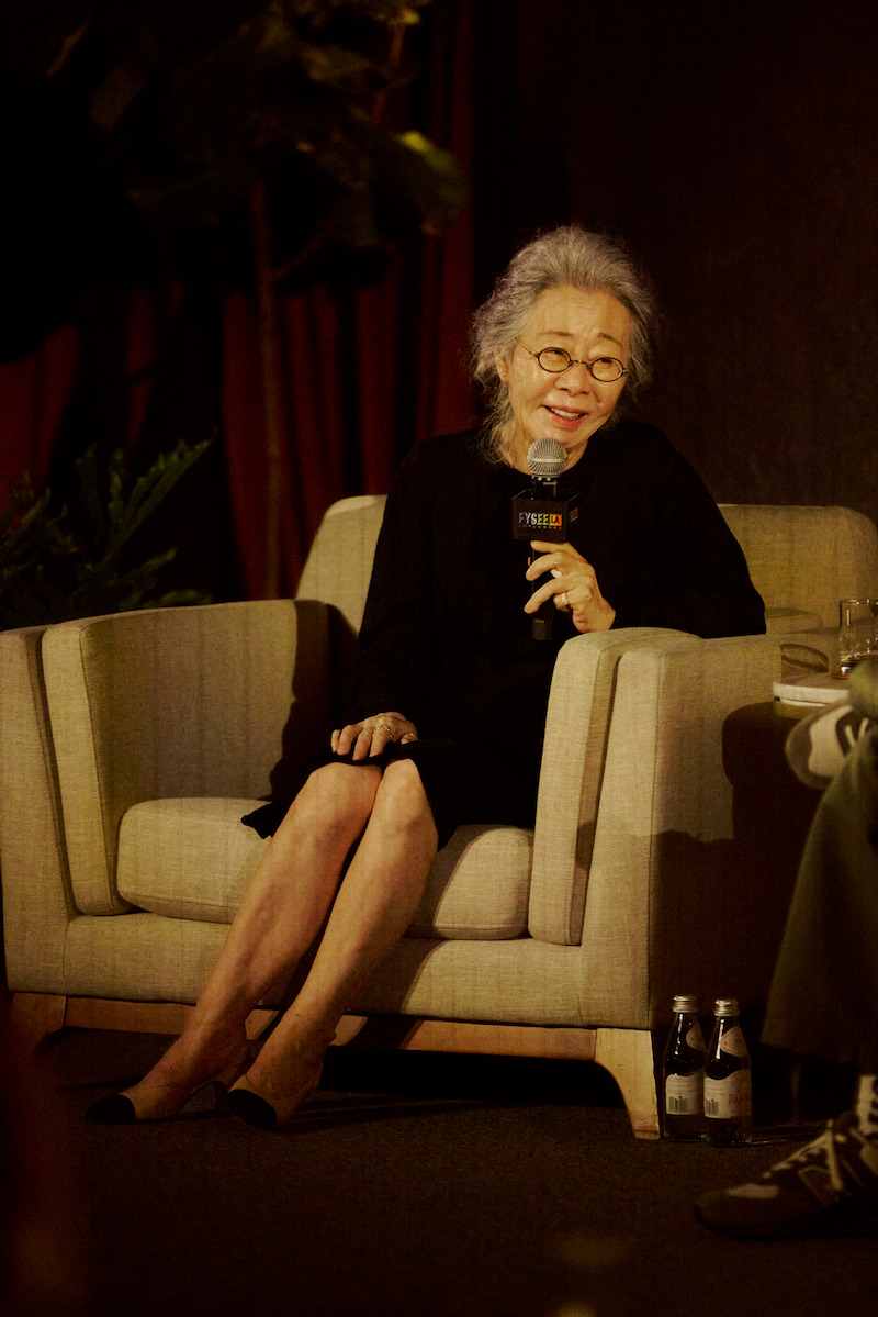 Elderly woman with gray hair and glasses sits on a beige armchair, speaking into a microphone at an indoor event, with plants and curtains in the background and water bottles on the floor.