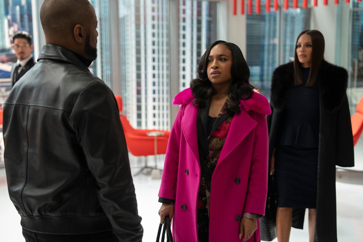 A woman in a bright pink coat stands facing a man in a leather jacket in a modern office with large windows and city views, while another woman in a dark outfit looks on.