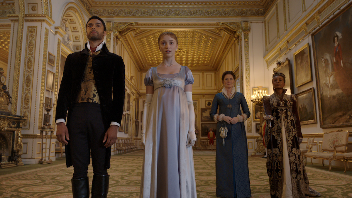 Four people in Regency-era attire stand in an ornate, gold-trimmed ballroom with chandeliers, patterned carpet, and large classical paintings on the walls. The setting suggests a formal or historic event.