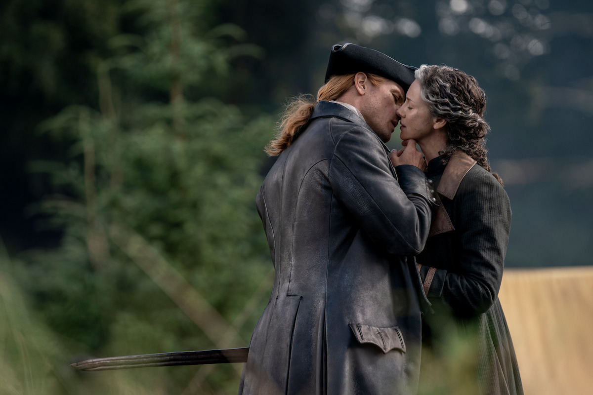 Two people in historical clothing share an intimate moment outdoors, surrounded by greenery. One wears a tricorn hat and long coat, the other a cape, suggesting a period drama or romantic scene set in nature.