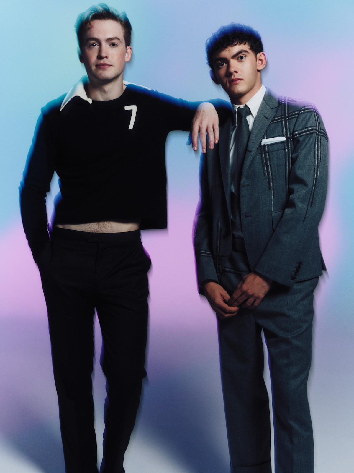 Two young men posing together in a studio with a soft pastel gradient background; one wears a black cropped sweater and pants, the other wears a gray suit, both looking at the camera with relaxed expressions.