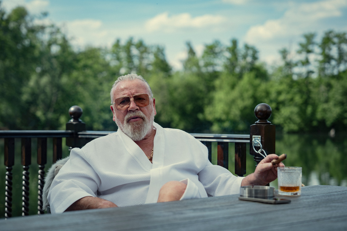 Older man in a white robe sits at an outdoor table by a lake, holding a cigar and a glass of whiskey, surrounded by greenery and partly cloudy skies, appearing relaxed in a natural, serene setting.
