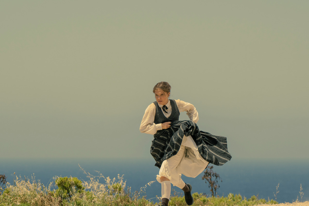 A woman in period clothing runs through a grassy landscape near the ocean under a clear, muted sky, suggesting a historical or dramatic outdoor scene with natural surroundings.