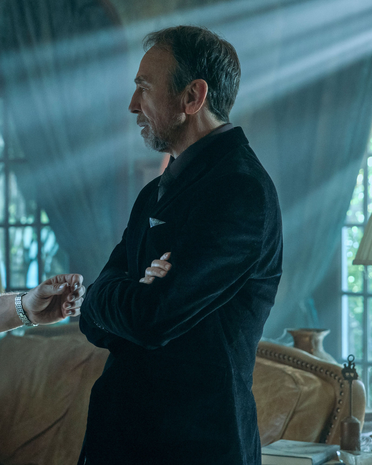 David Jensen as Wes Genrette with beard in dark suit standing with arms crossed in a dimly lit elegant room, with sunlight streaming through large windows and draped curtains, conversing with someone partially visible on the left.
