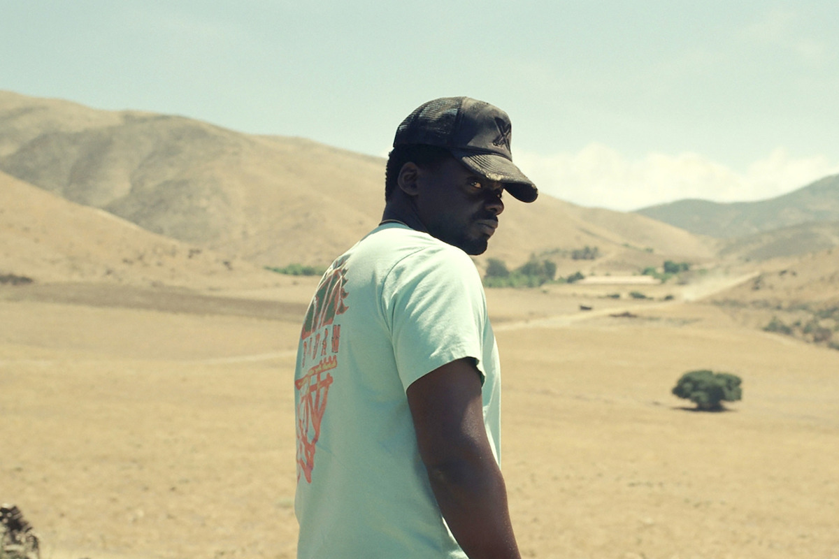 Man wearing a cap and sunglasses stands outdoors in a sunlit, dry, hilly landscape with a winding road and scattered trees under a partly cloudy sky.