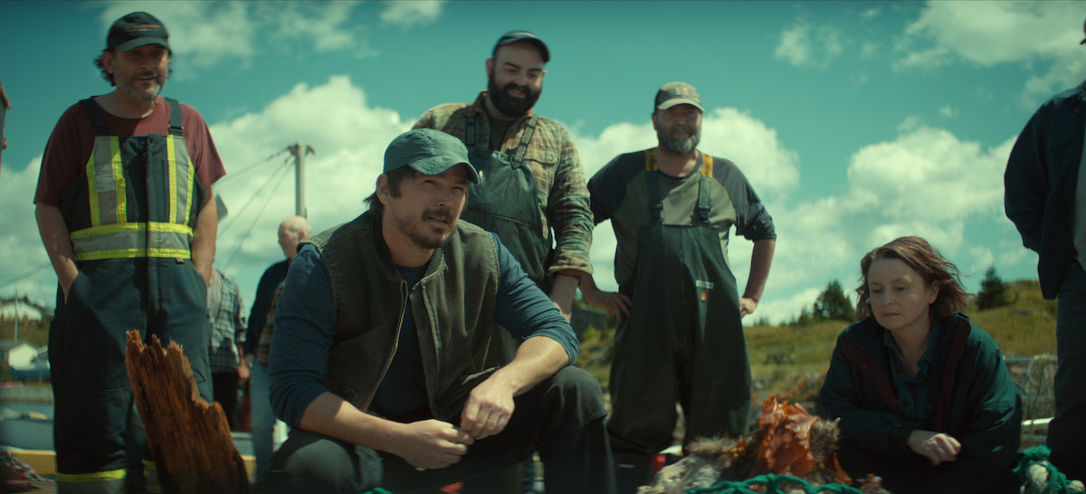 A group of fishermen and a woman crouch and stand outdoors near fishing gear under a bright blue sky with clouds, on a grassy landscape possibly near the sea.