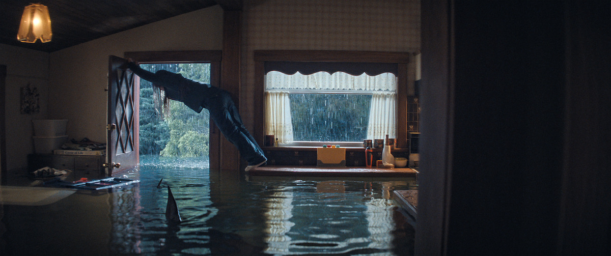 Person in flooded house opening door, heavy rain visible outside, shark fin in water, kitchen in background, indoor lighting, dramatic and surreal atmosphere.