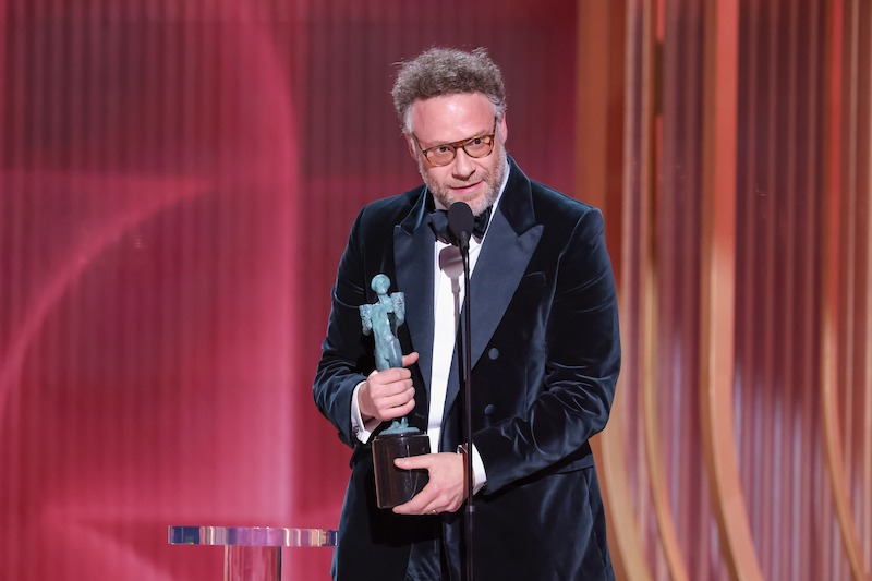 Seth Rogen holding a trophy and speaking into a microphone on stage with a red and orange backdrop, likely at an awards ceremony.