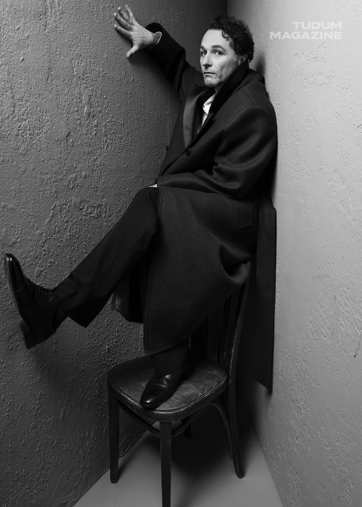Matthew Rhys poses for the camera wearing all black standing on a wooden chair.
