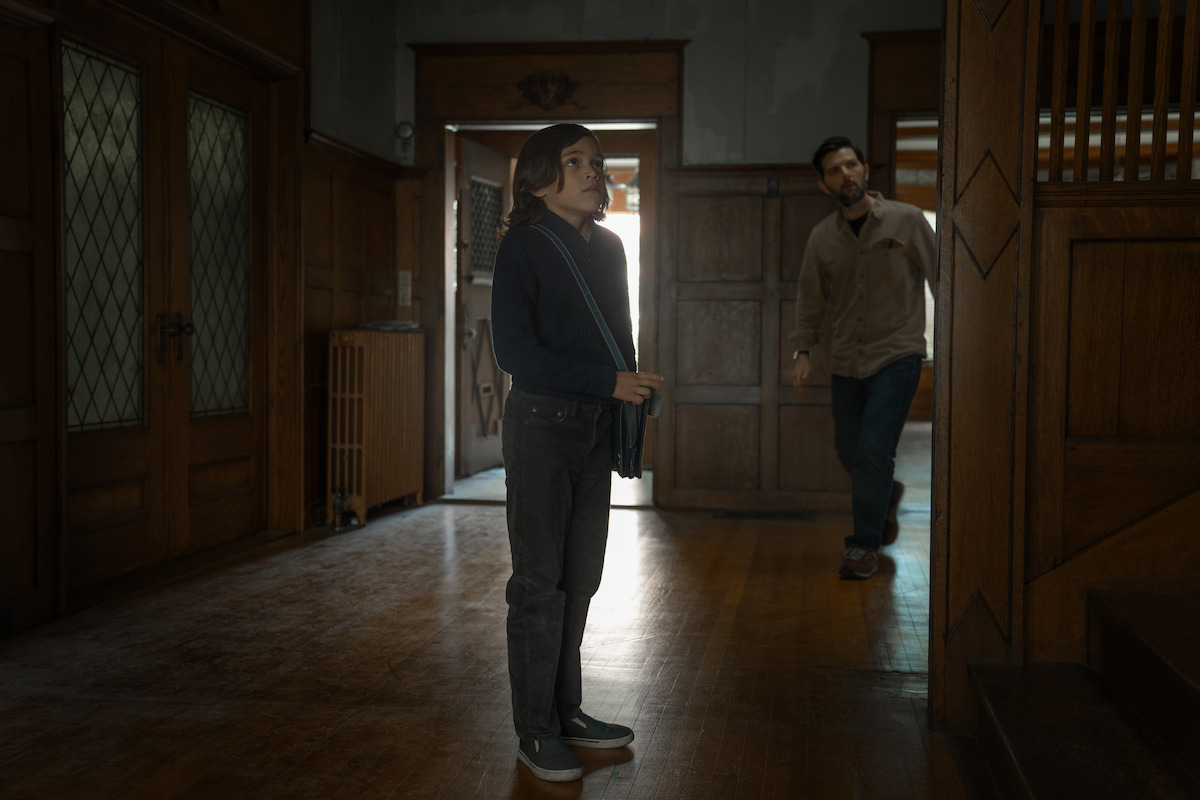 A woman stands in a spacious, dimly lit room with wood paneling, while a man enters from a doorway. The setting appears to be an old house or building with vintage architecture and wooden floors.
