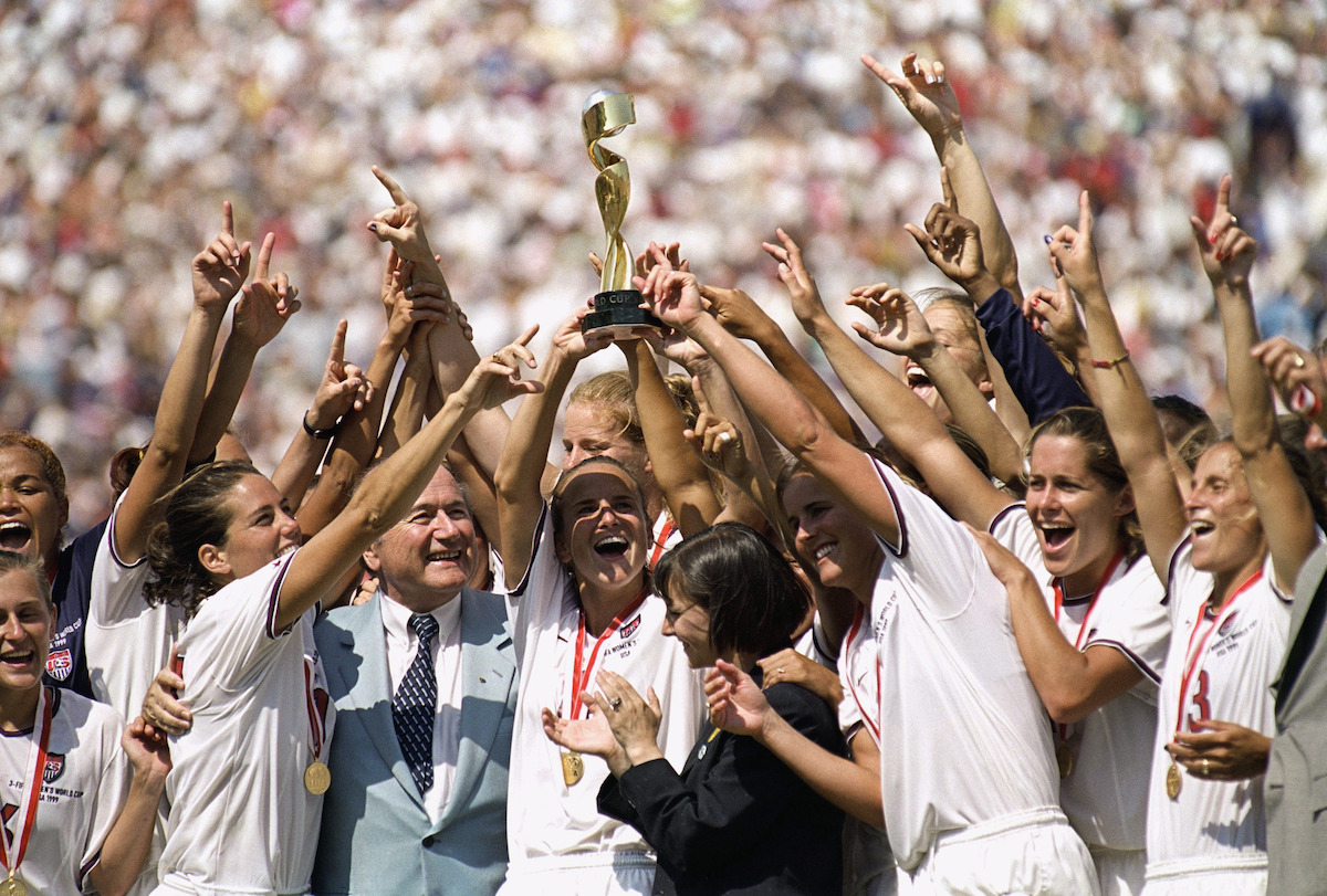 Soccer team in white uniforms celebrates together, holding a trophy high, with medals around their necks. Crowd of fans in the background; outdoor stadium setting, daytime, joyful victory moment.