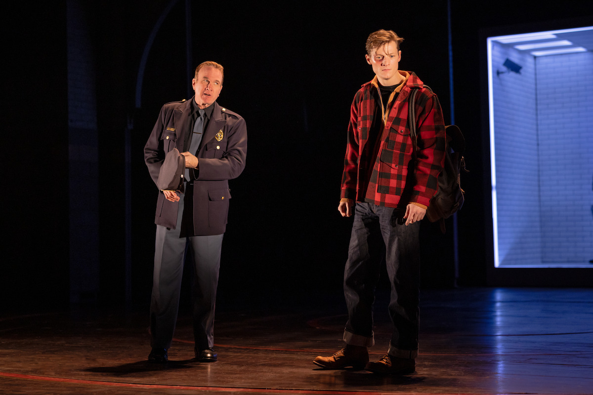 A police officer and a young man in casual clothes stand on a dimly lit stage with a blue-lit modern room set in the background, suggesting a dramatic theater scene.