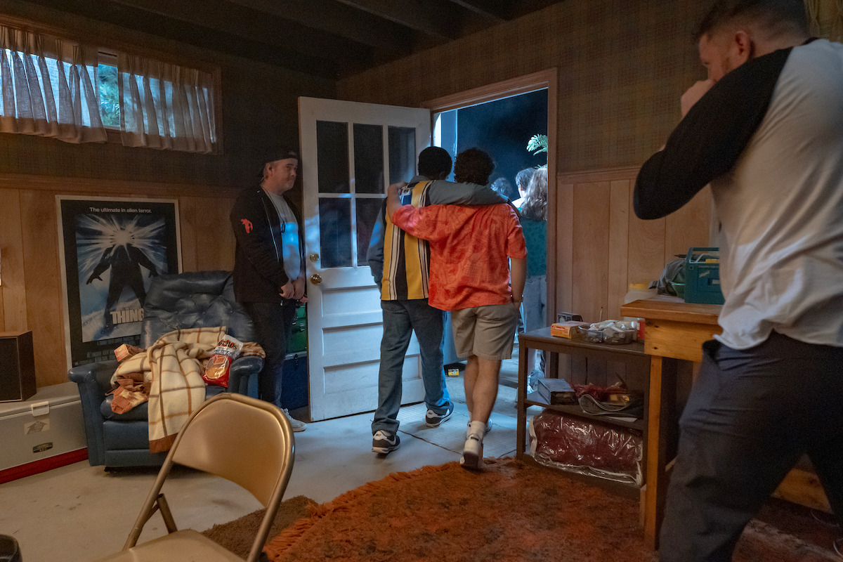 Two friends with arms around each other exit a cozy, cluttered room through an open door.