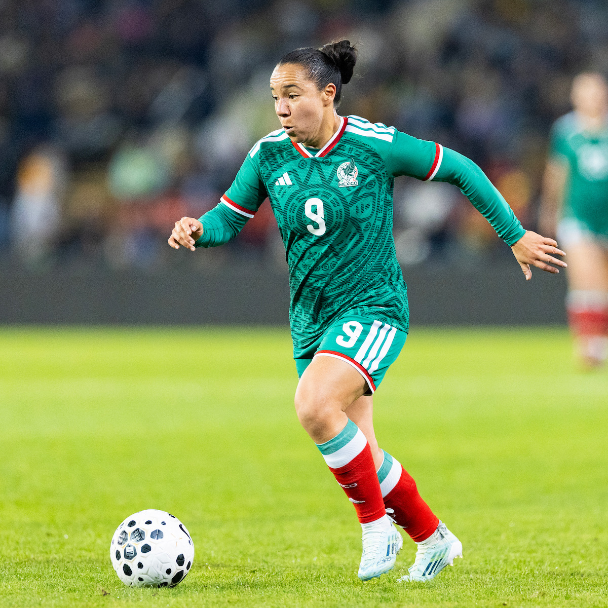 Soccer player in green Mexico jersey dribbles a ball on a stadium field during a match, with blurred crowd and other players in the background.