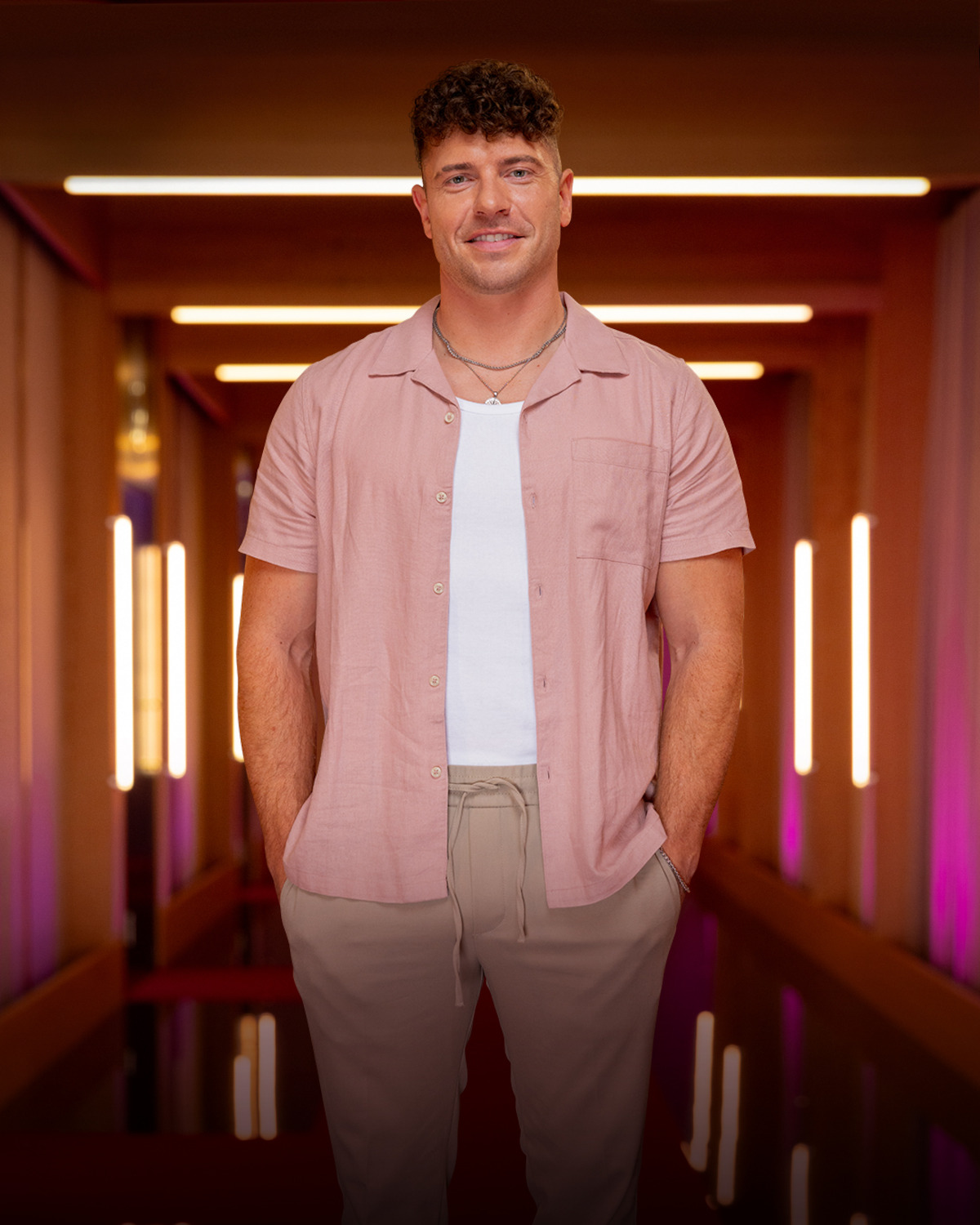 Man with short curly hair in casual pink shirt and beige pants stands in a modern, warmly lit hallway with neon vertical lights and pink accents, facing the camera with hands in pockets.