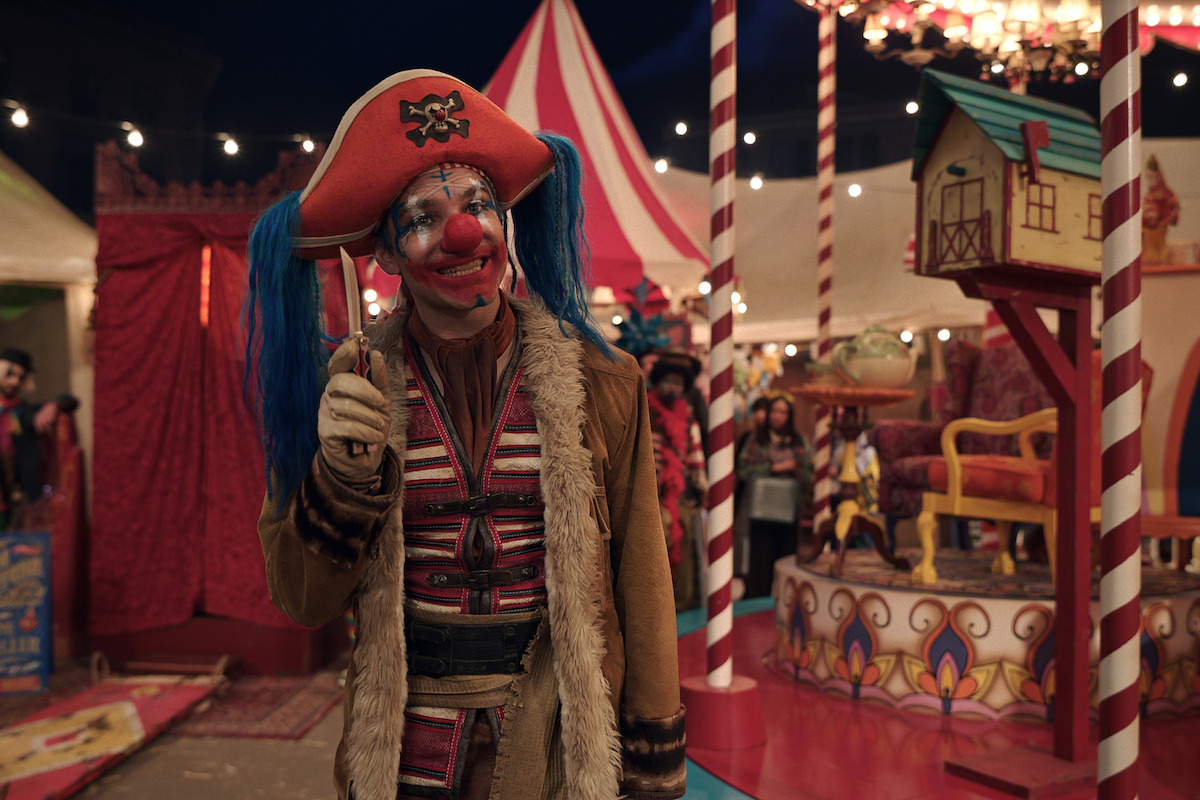 A clown in a red pirate hat, blue pigtails, and colorful costume stands smiling in a lively circus setting with striped tents, bright lights, festive decor, and people in the background.