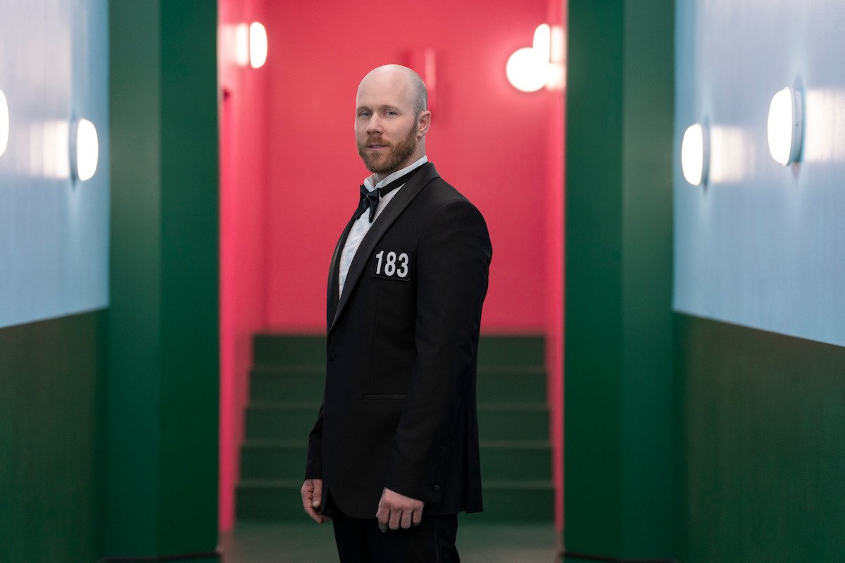 A man in a formal black suit stands in a brightly lit hallway with blue and green walls and a pink background, giving a dramatic and tense atmosphere. The number 183 is on his jacket.