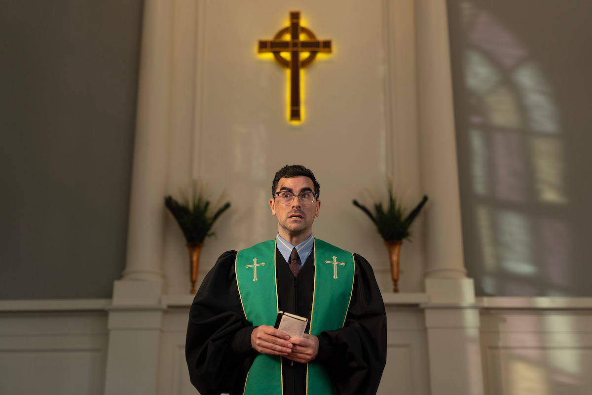 A pastor in black and green robes stands holding a book in a church sanctuary, with a large illuminated cross and plants behind him, soft light coming through stained-glass windows.