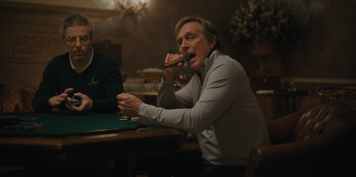 Two men at a poker table in a dimly lit, elegant room; one shuffles cards while the other bites a cigar and holds poker chips, with decorative flowers and ornate furnishings in the background.